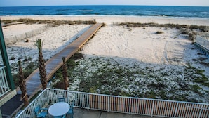 Een uitzicht op het strand met een houten vogelzangpad dat naar de oceaan leidt, een omheind gebied met stoelen en een gebouw met een balkon.