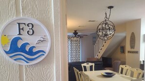 A dining area with a white table and chairs, a chandelier, and a wall-mounted sign with 'F3' and a wave design. - Beautiful 3 Bedroom BEACHFRONT Condo - Sundial #F3 - Sleeps 10 (Gulf Shores)