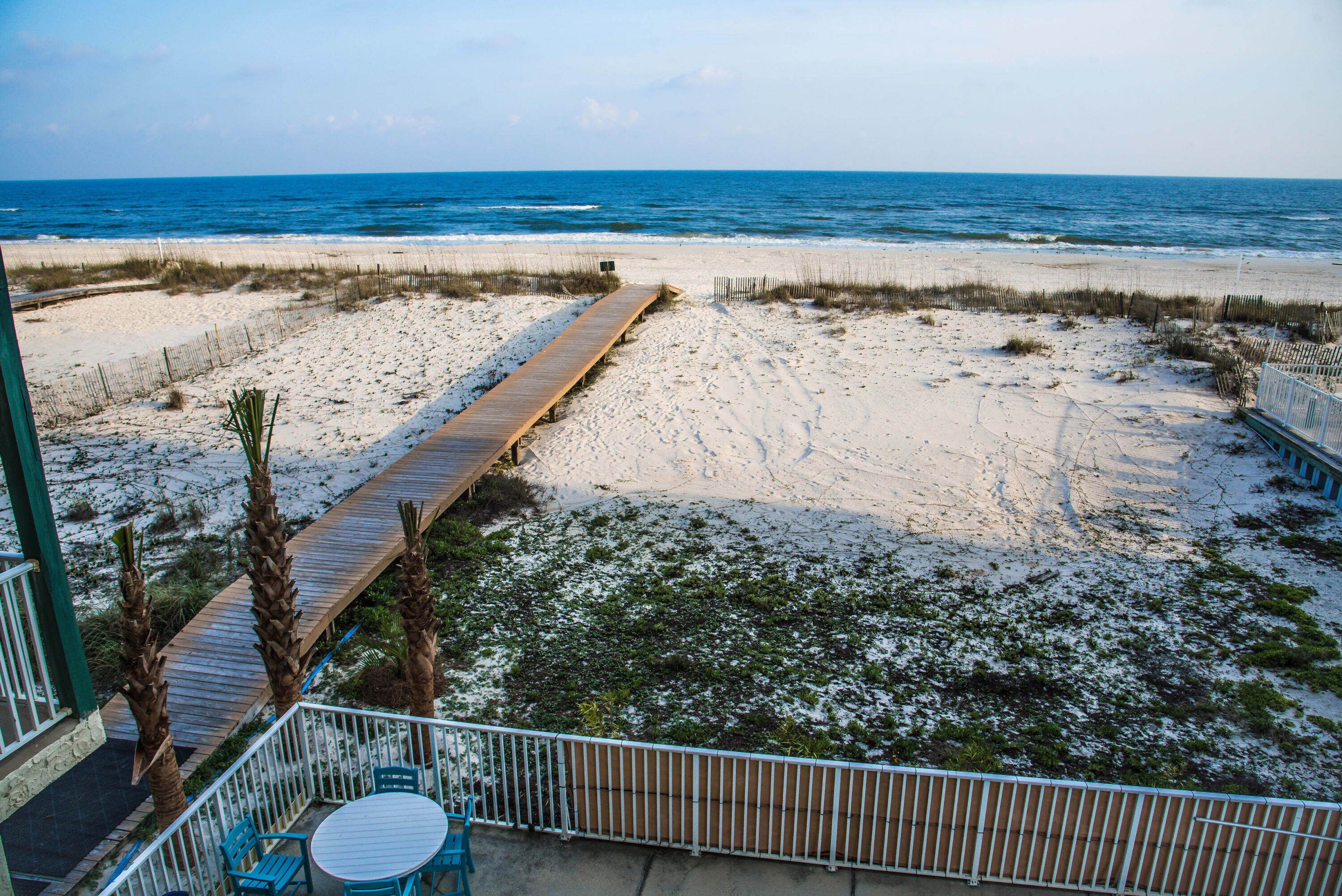 Vista de la playa con un paseo marítimo de madera que conduce al océano, un área cercada con sillas y un edificio con balcón.