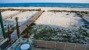 A beach view with a wooden boardwalk leading to the ocean, a fenced area with chairs, and a building with a balcony. - Beautiful 3 Bedroom BEACHFRONT Condo - Sundial #F3 - Sleeps 10 (Gulf Shores)