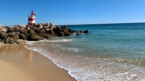 Uma praia com um farol numa formação rochosa, céu azul límpido e mar calmo.