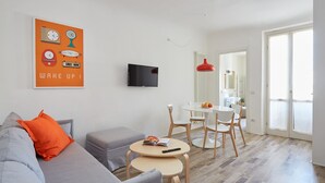 A modern living room with a grey sofa, a small round table, a TV mounted on the wall, and a dining area with a white table and chairs.