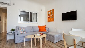 A modern living room with a grey sofa, a round coffee table, a TV mounted on the wall, and a dining area with a white table and chairs.