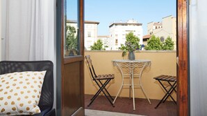 Un balcon donnant sur des immeubles et une église, une chaise en osier, une petite table et un coussin.