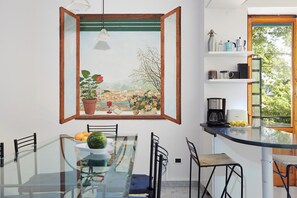 Uma sala de jantar com mesa de vidro, cadeiras, planta em vaso e vista para a cidade pela janela.