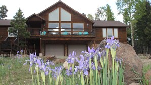 Una cabaña de madera con balcón, rodeada de paisaje natural y flores moradas.