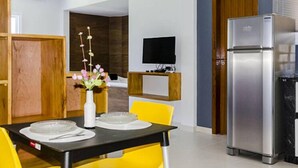 Une cuisine moderne avec un réfrigérateur en acier inoxydable, une table à manger avec des chaises jaunes et un téléviseur à écran plat fixé au mur.