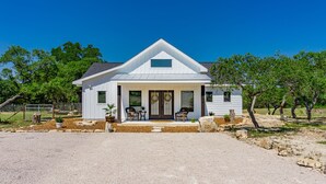 Une maison blanche d’un seul étage, avec un toit à pignon, un porche à l’avant doté de deux chaises et un petit jardin.