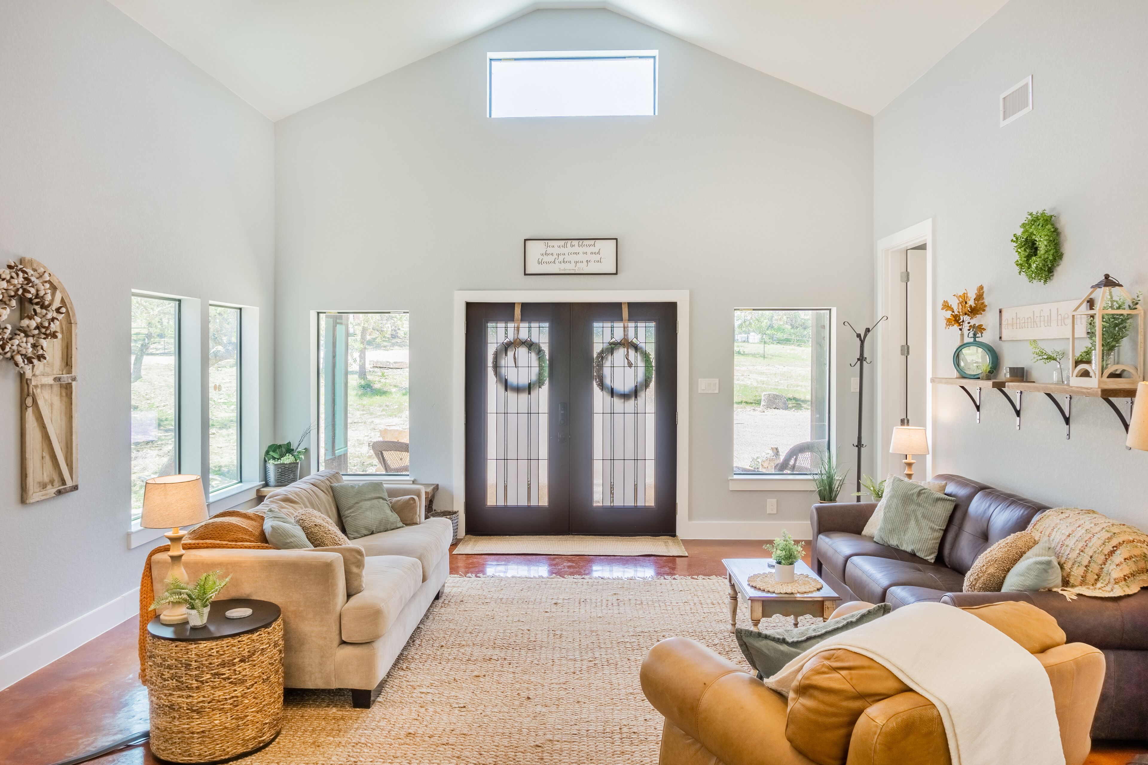 A spacious living room with a high ceiling, a large window, a dark front door with decorative wreaths, and a cozy seating area with a brown leather sofa and a beige couch.