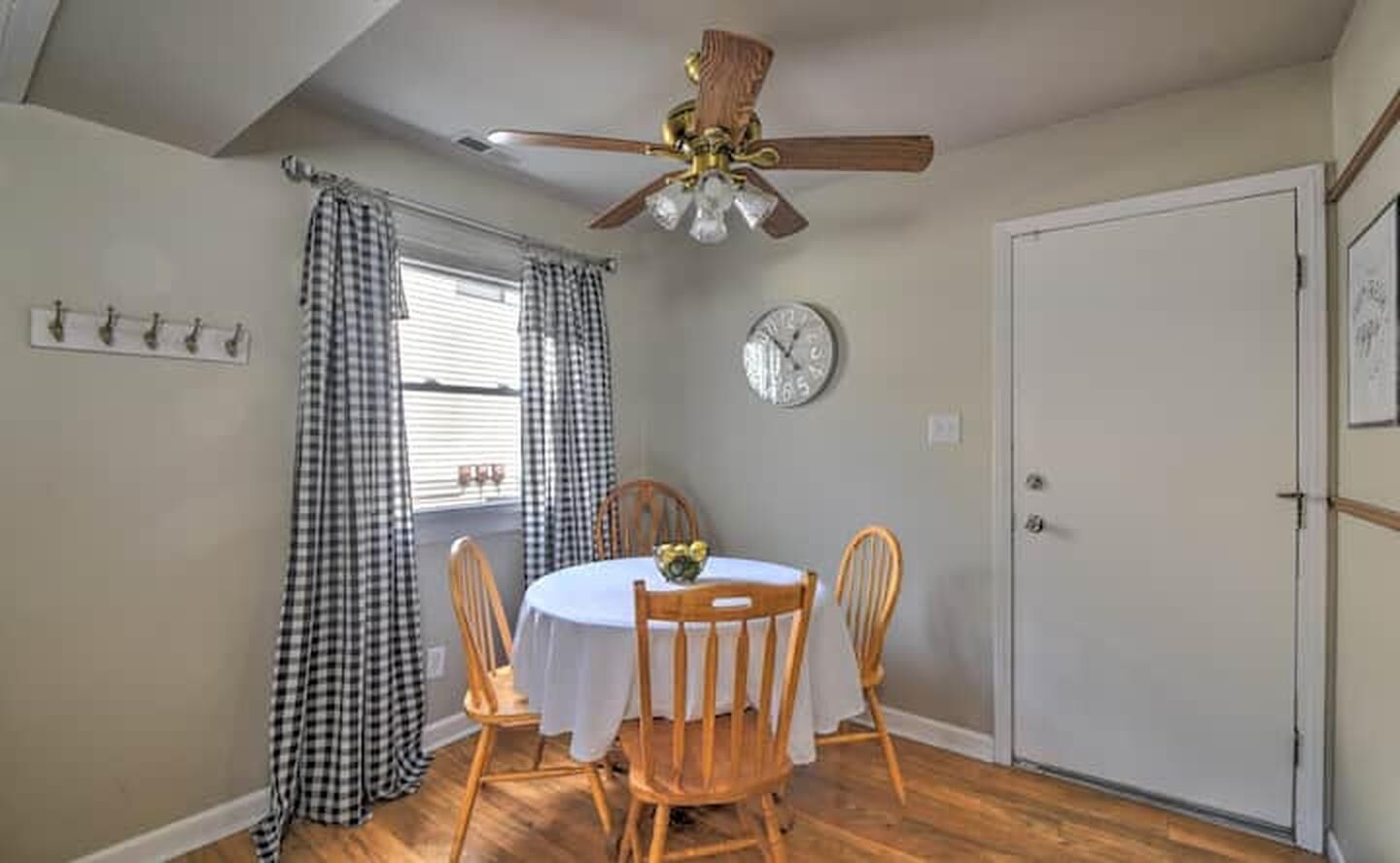 A dining area with a round table, four chairs, a ceiling fan, a clock, and a door.