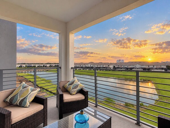 A balcony with wicker furniture and a view of a sunset over a body of water and buildings.