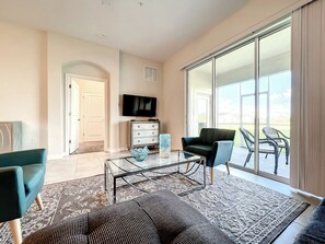 A spacious living room with a large sliding glass door leading to an outdoor area, a flat-screen TV mounted on the wall, and a coffee table with decorative items.