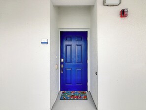 Una puerta azul con una placa de número, un felpudo y un detector de incendios en la pared.