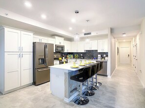 Cocina moderna con armarios blancos, electrodomésticos de acero inoxidable, una isla central con taburetes y luminarias colgantes.