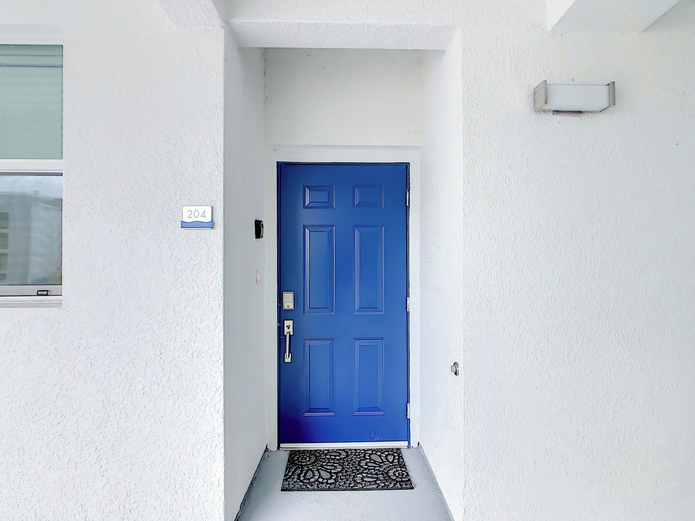 Una puerta azul con una placa de número, un ventanal pequeño y un timbre en una pared blanca.