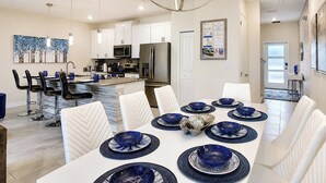 Una cocina moderna con armarios blancos, electrodomésticos de acero inoxidable y una zona de comedor con una mesa blanca para seis personas.