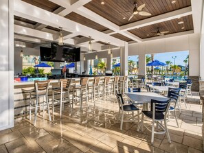 An outdoor patio area with seating and a bar, featuring a pool view and ceiling fans.