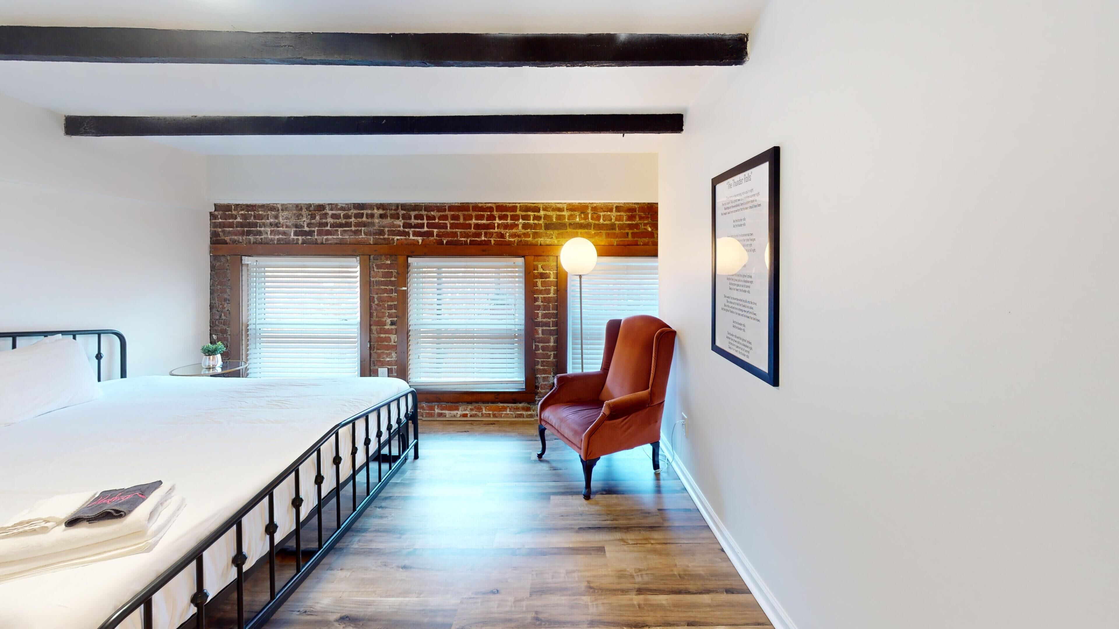 Een slaapkamer met een bed, een stoel, een bakstenen muur en een raam met jaloezieën.