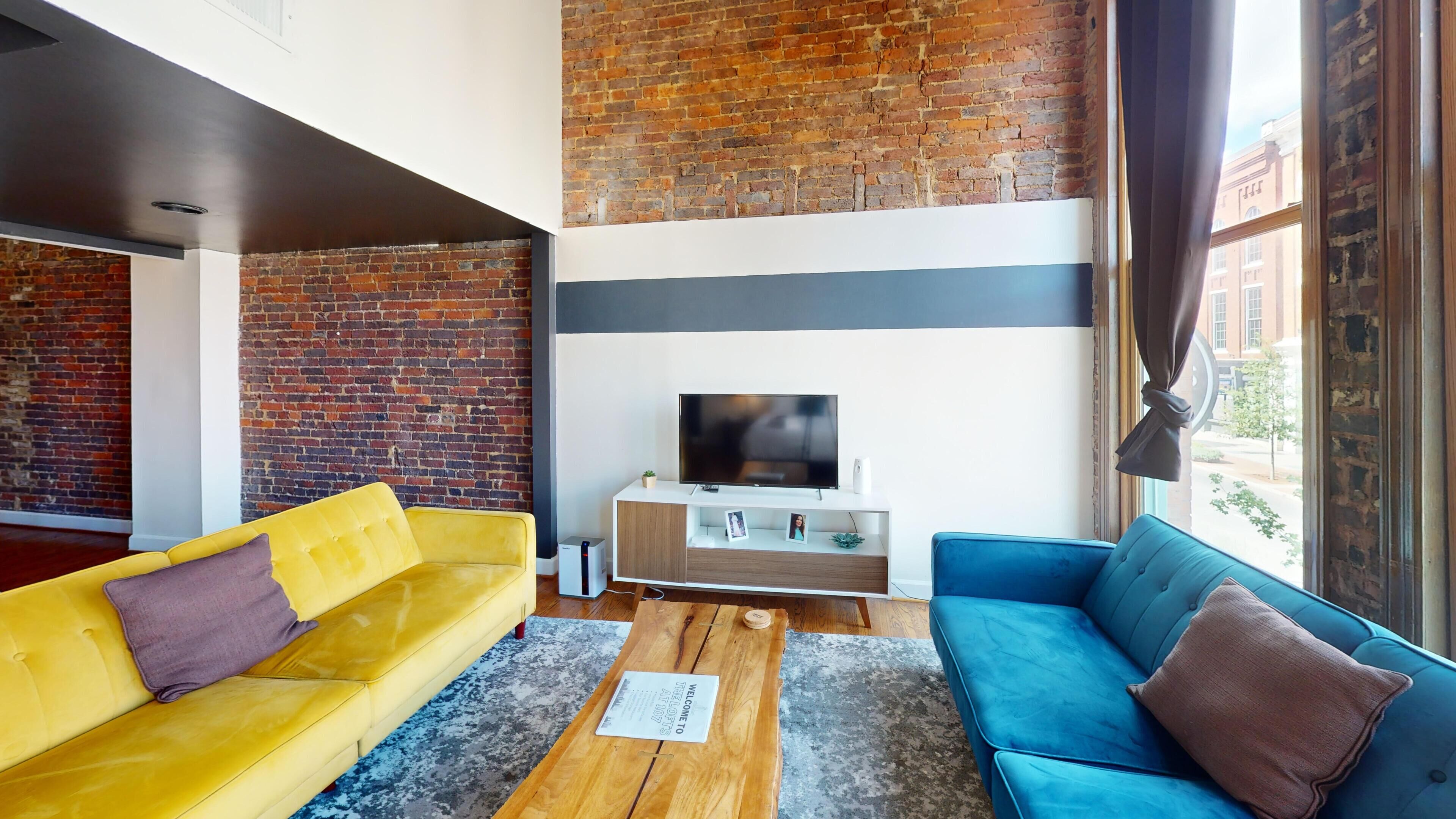 Una sala de estar moderna con pared de ladrillo, televisor de pantalla plana, sofás amarillos y azules, y una mesa de centro de madera.
