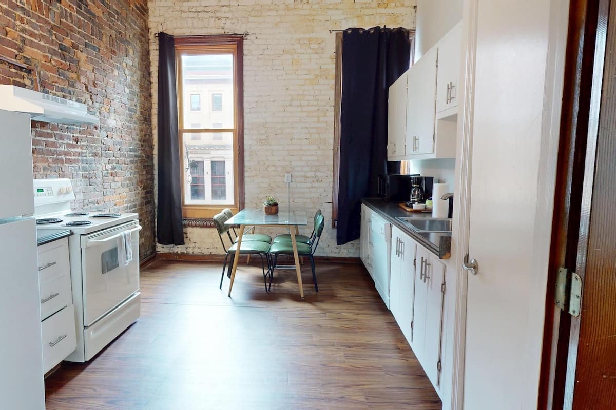 A compact kitchen with a brick wall, a window with curtains, a dining table with chairs, and white cabinetry.