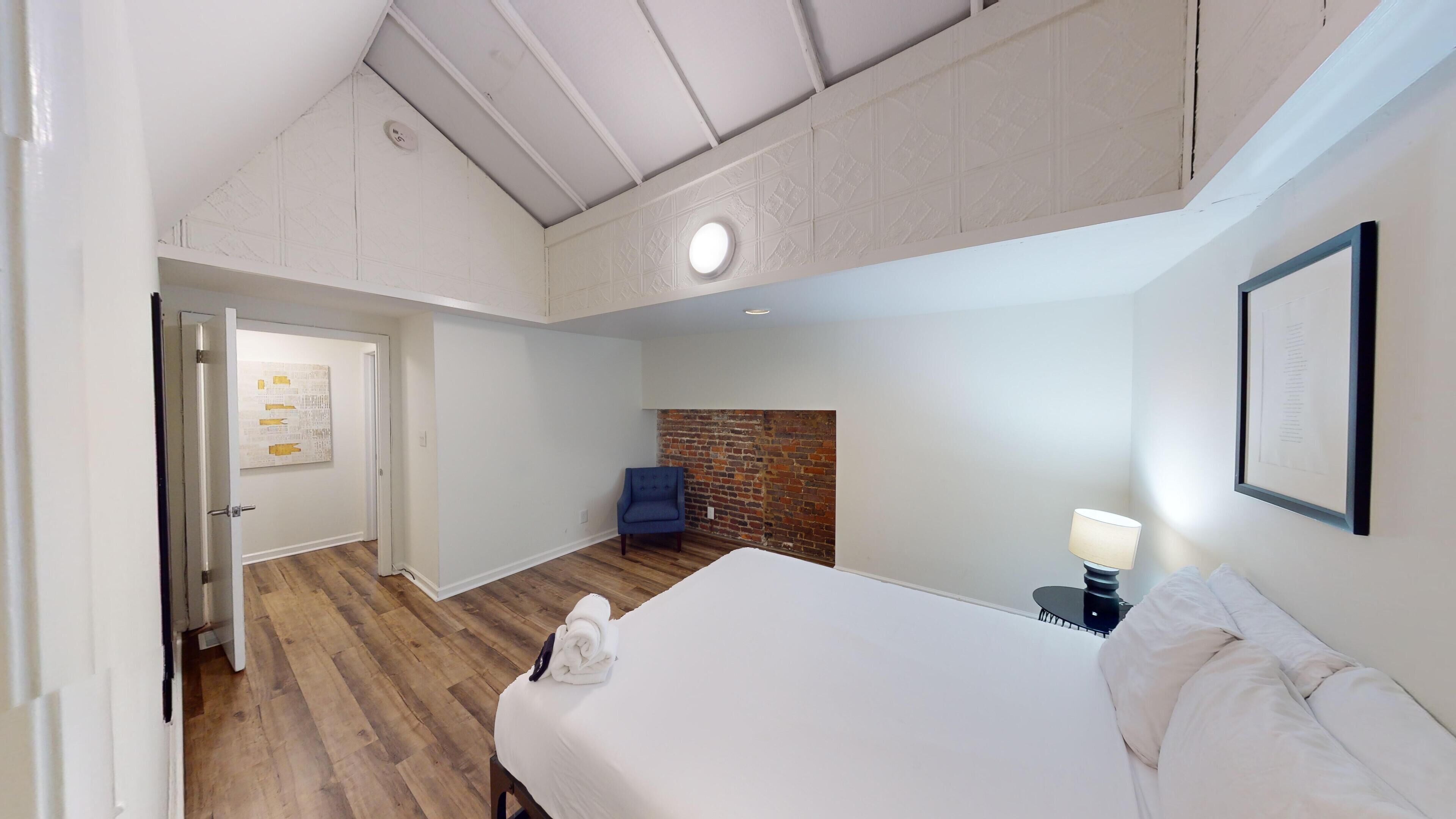 A bedroom with a bed, a blue chair, a brick wall, and a white door with a poster.