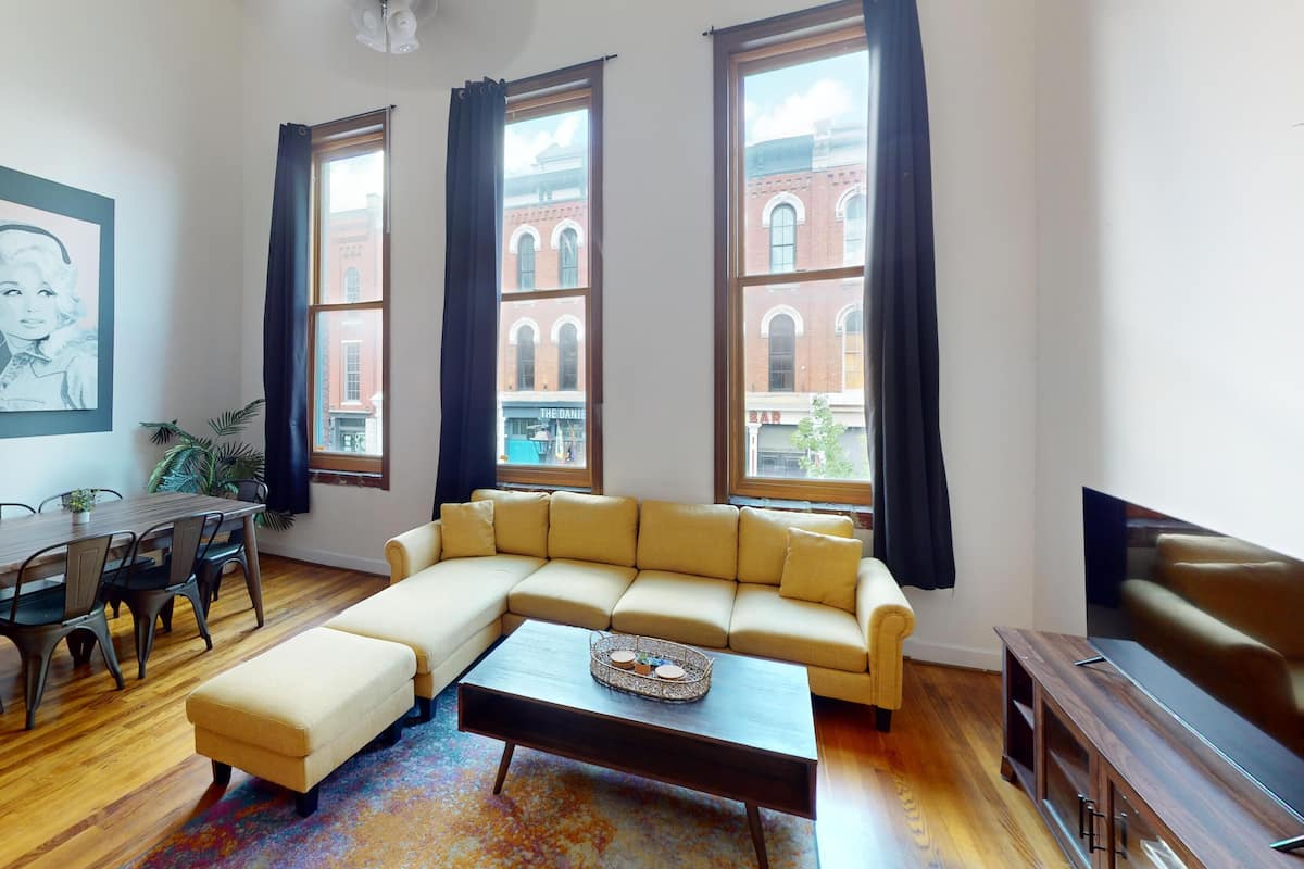 A living room with a sofa, a coffee table, a dining table with chairs, and large windows with curtains.