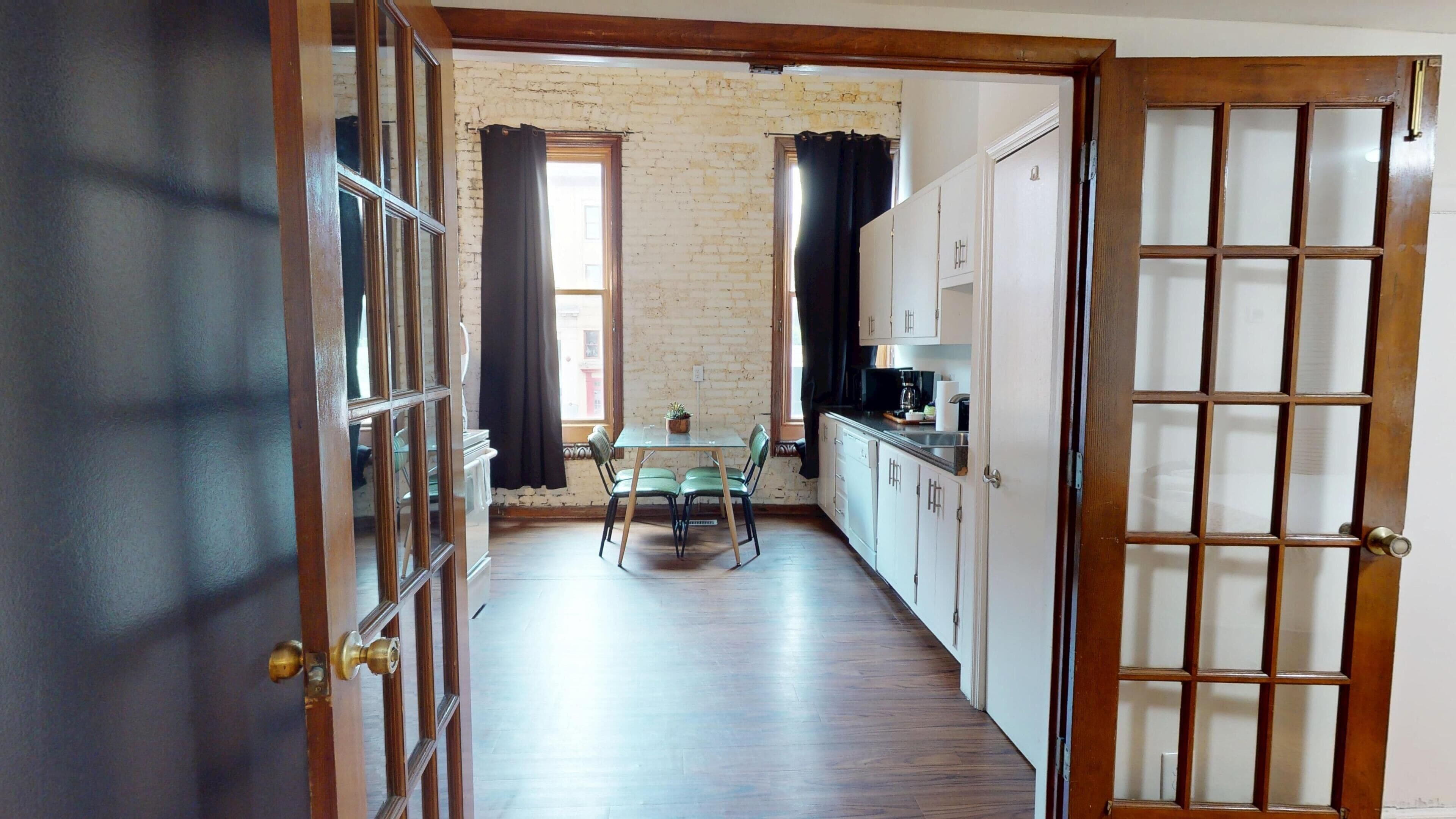 A narrow hallway leading to a dining area with a table and chairs, wooden flooring, and a brick wall.