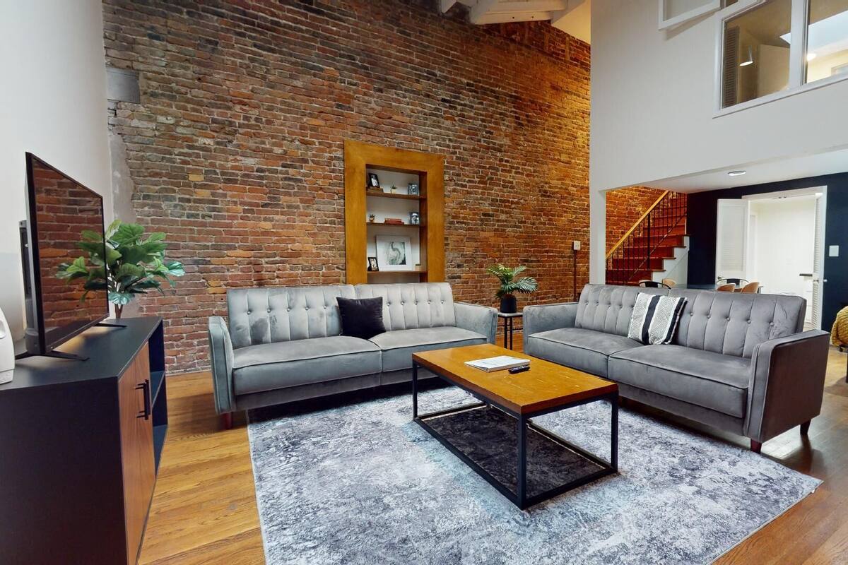 A modern living room with a brick wall, grey sofas, a wooden coffee table, and a staircase leading to another level.