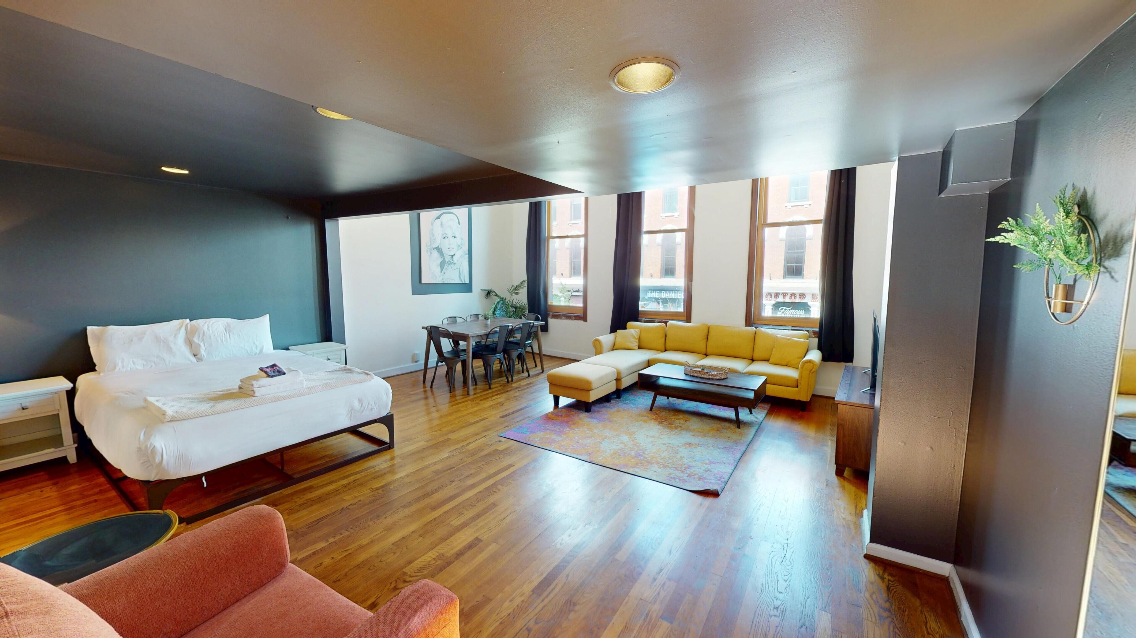 Un salon moderne comprenant un lit, un canapé jaune, un fauteuil rose et un coin repas avec une table et des chaises.