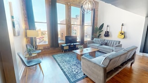 Una sala de estar moderna con un sofá gris, una mesa de centro y un televisor. Hay dos guitarras en la pared, una planta y un ventanal con cortinas.