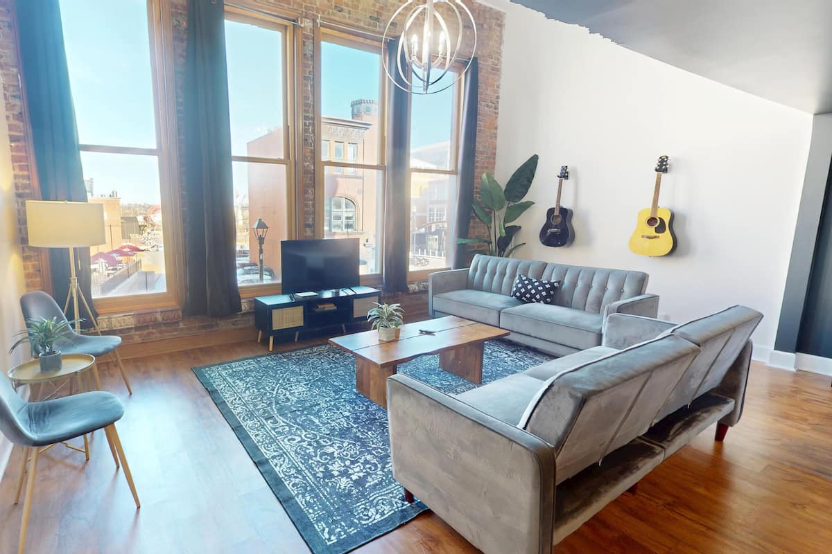 A modern living room with a grey sofa, a coffee table, and a TV. There are two guitars on the wall, a plant, and a large window with curtains.