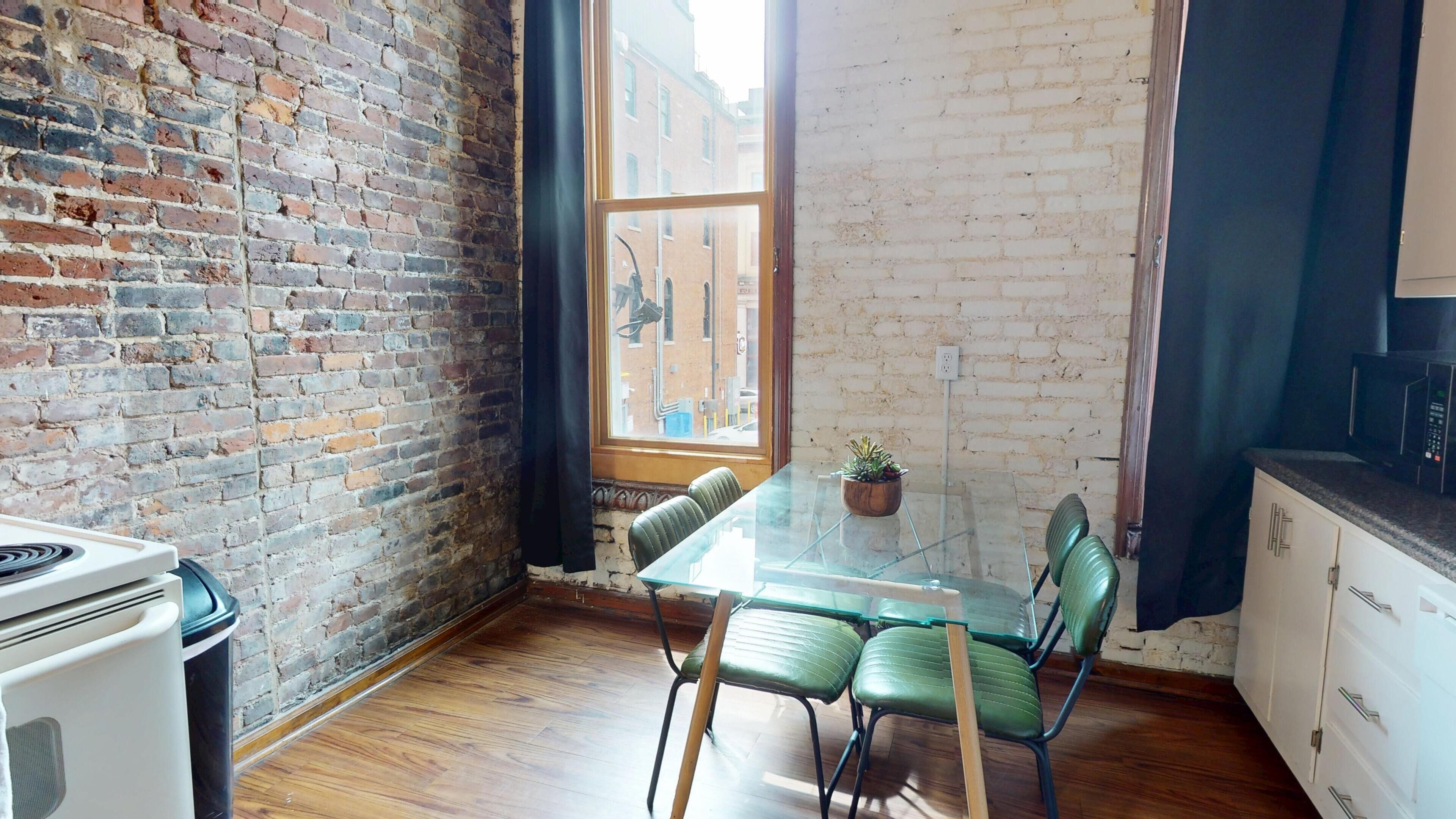 Una cocina con una mesa de vidrio, sillas verdes, un muro de ladrillo y una ventana.