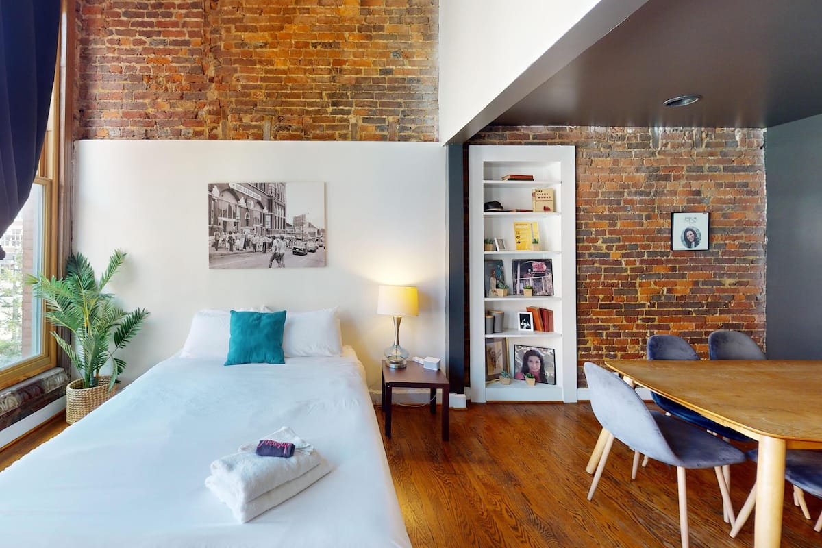A hotel room with a bed, a bedside table, a lamp, a bookshelf, and a dining area with chairs.