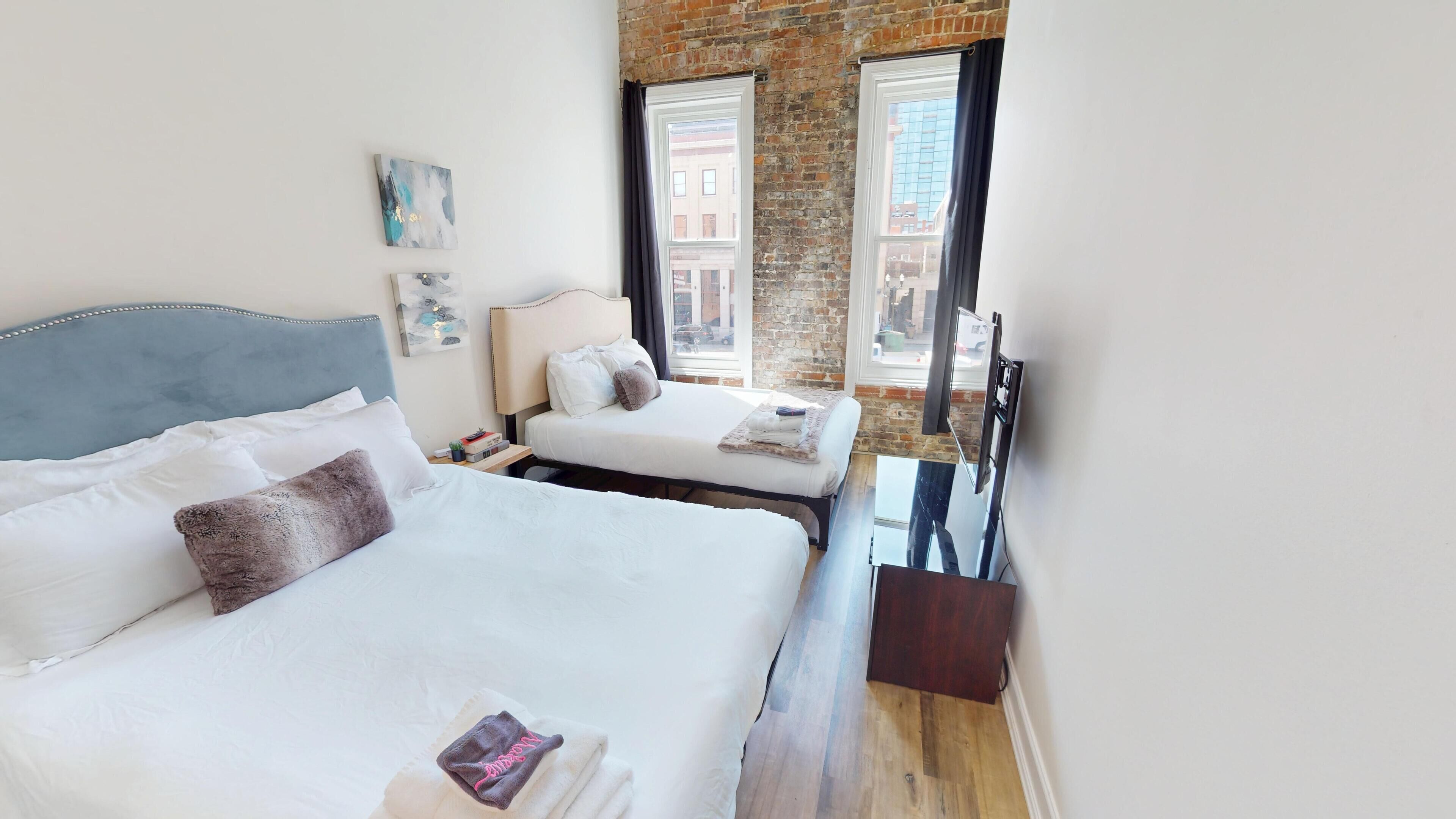 Habitación de hotel con dos camas, una pared de ladrillo y ventanas grandes.