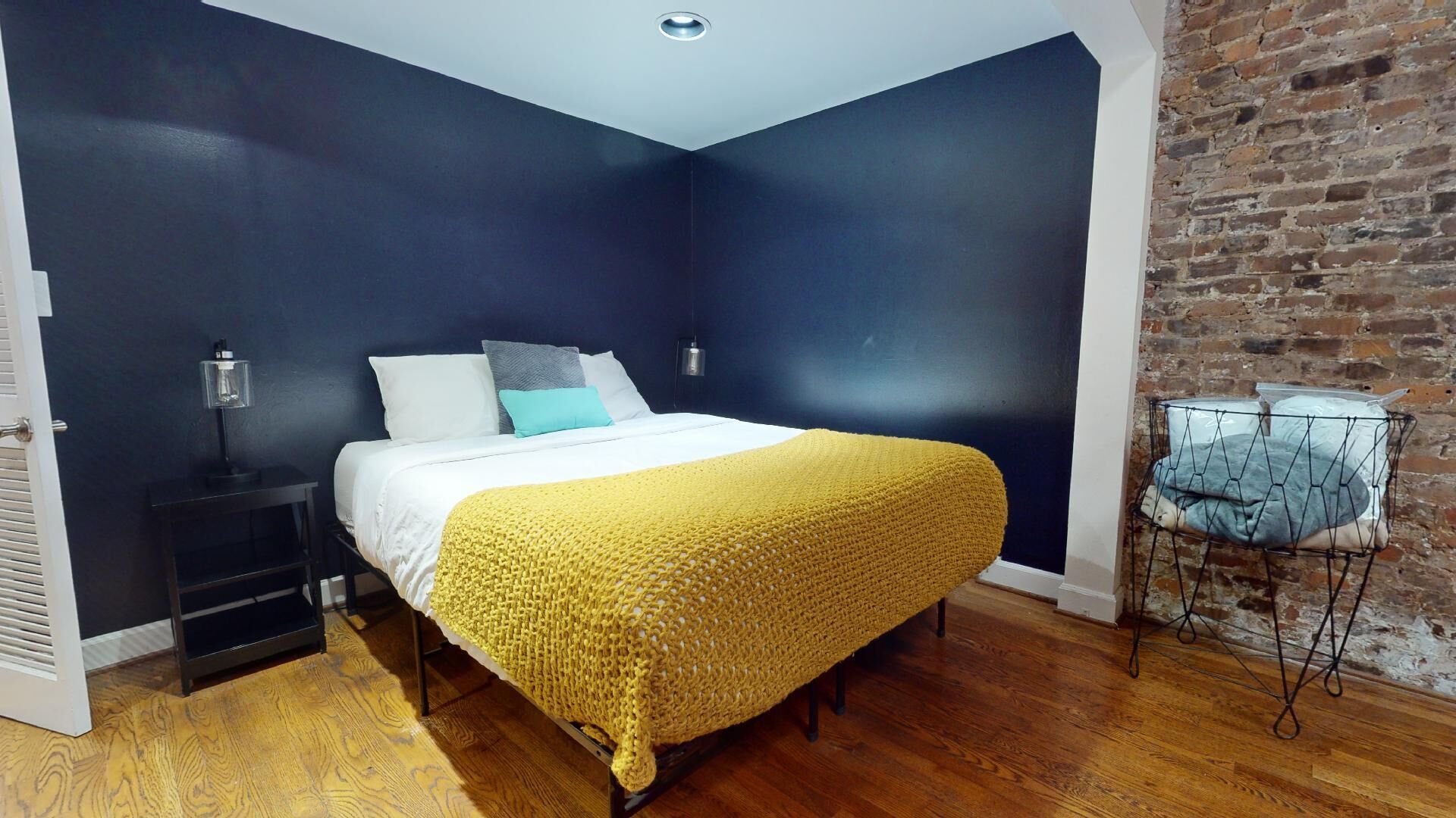 A bedroom with a bed, a nightstand, a lamp, a brick wall, and a metal chair.