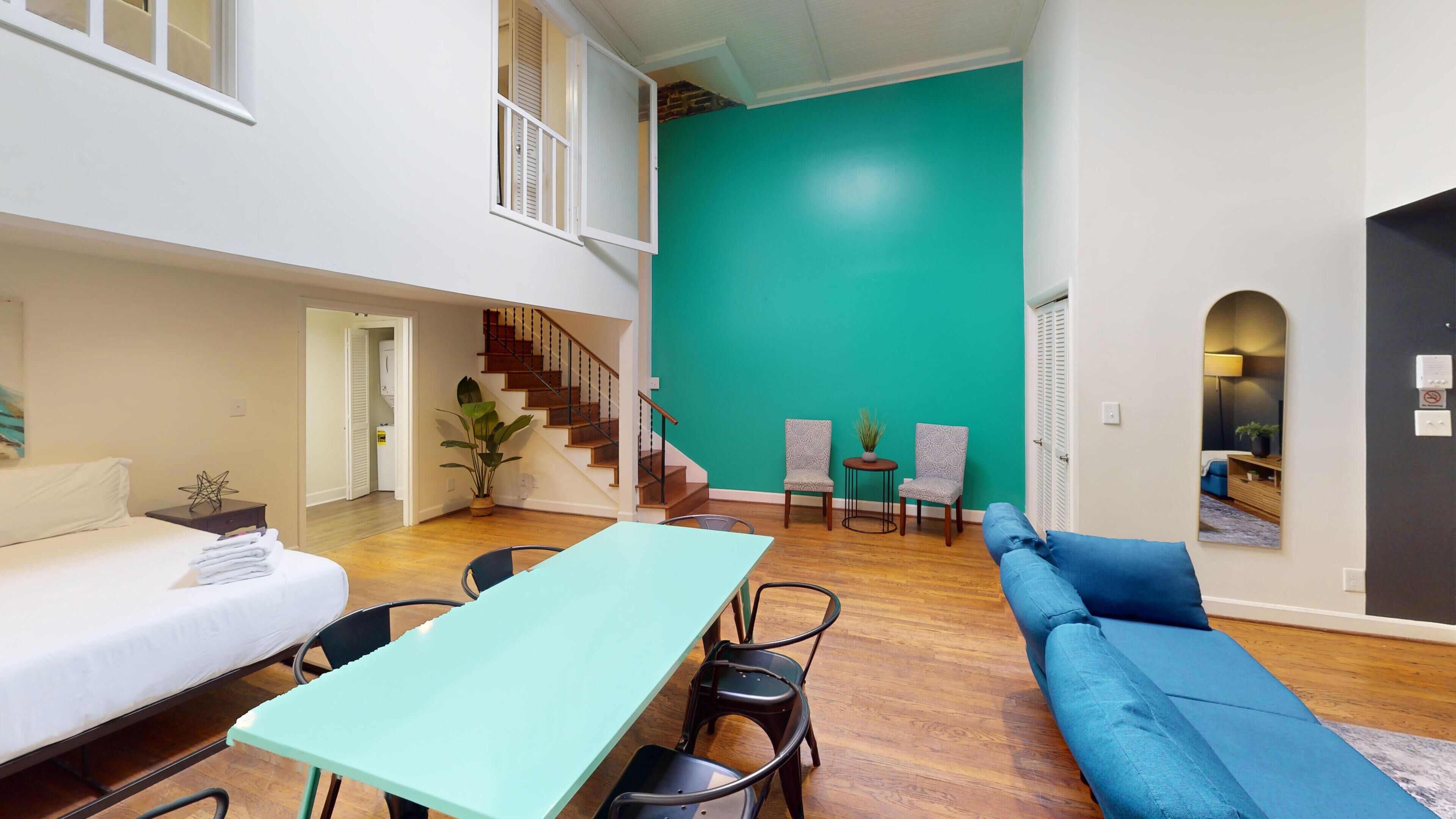 Amplia sala de estar con una pared verde azulado, una escalera, una mesa de comedor con sillas, un sofá y una pequeña mesita de noche con una planta.