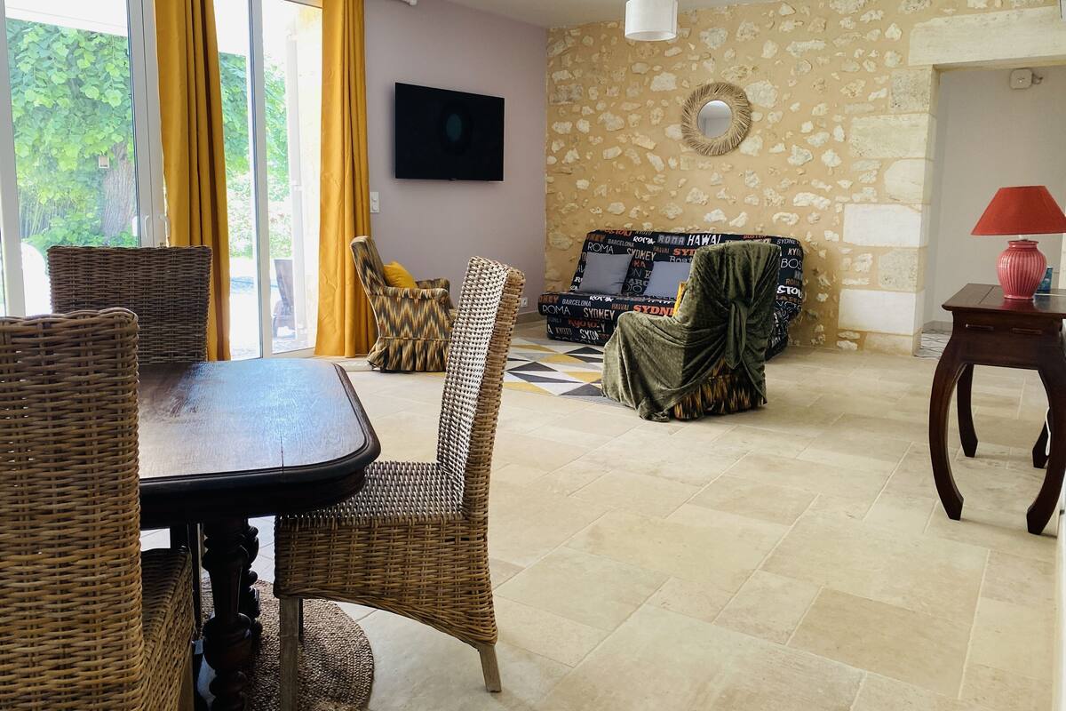 Un salon comprenant une table à manger, des chaises en osier, un canapé avec des coussins à motifs, un téléviseur et un mur en pierre orné d’un miroir rond.
