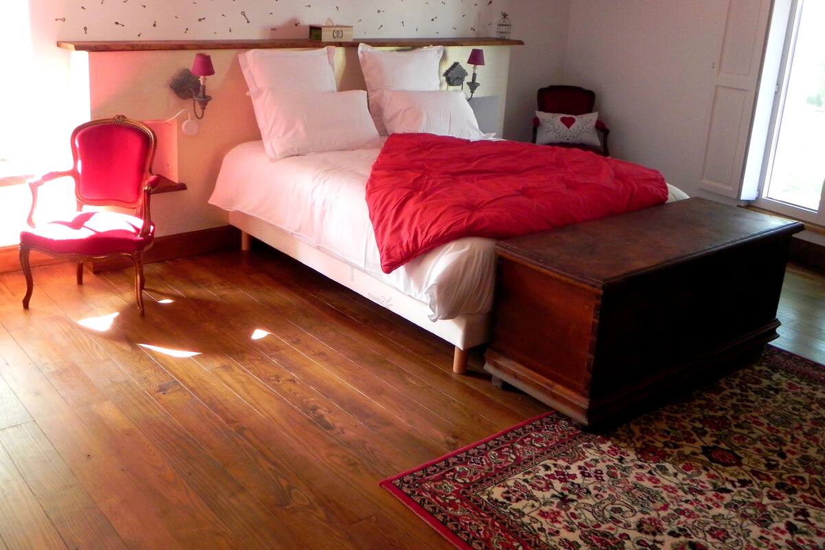 Une chambre à coucher avec un lit, une chaise rouge, une commode en bois, un tapis à motifs et un mur agrémenté de quelques éléments décoratifs.