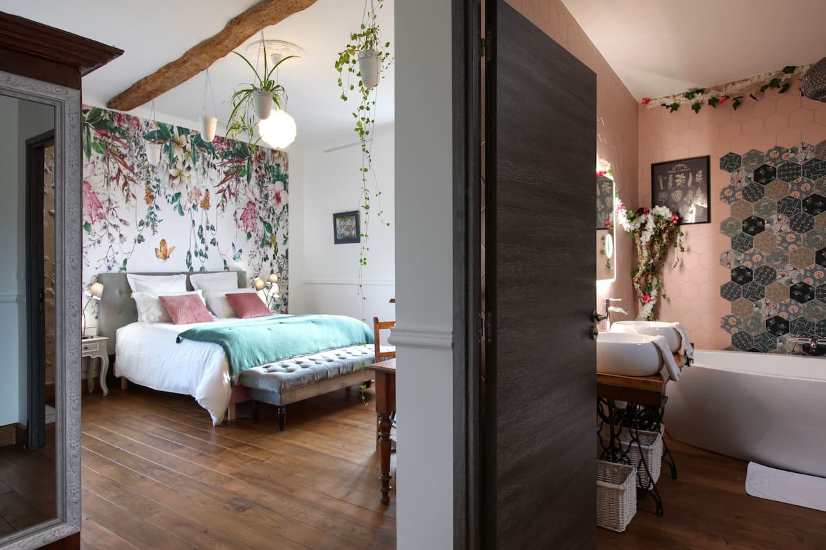 Une chambre à coucher avec un lit, une table de chevet et une commode. On y trouve une fenêtre avec des rideaux, un mur orné d’un motif floral et une poutre en bois au plafond.