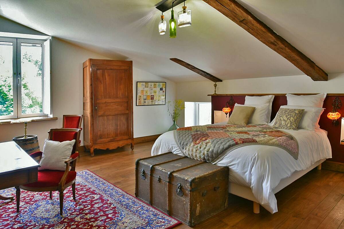 Une chambre avec un grand lit, des poutres apparentes en bois au plafond, une vieille malle et un tapis aux motifs rouges et bleus.