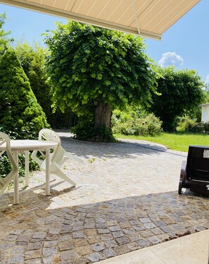 Une terrasse dallée avec une table et des chaises en plastique blanc, un grand arbre au feuillage dense et un ciel bleu clair.