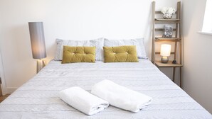 A neatly made bed with two pillows, a bedside table with a lamp, and a small shelf with decorative items. - Sunshine Place (Enfield)