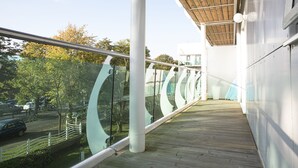 A modern building with a glass railing, a wooden deck, and a view of a parking area and trees. - Sunshine Place (Enfield)