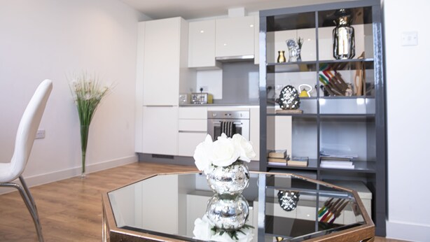 A modern kitchen with white cabinets, a glass-top dining table, and a vase with white flowers. - Sunshine Place (Enfield)