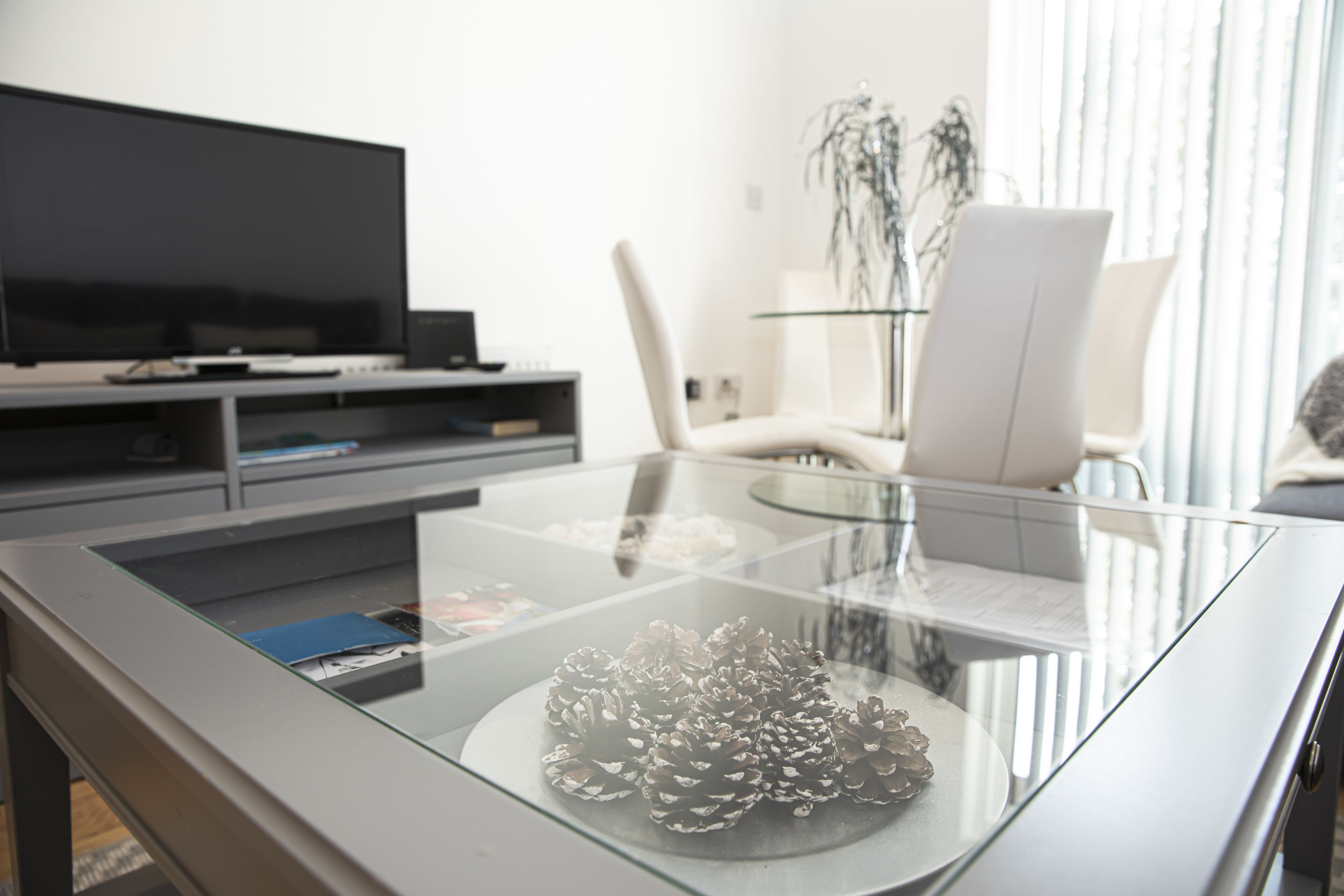 A modern living room with a glass coffee table, a flat-screen TV, a white chair, and a plant.