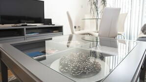 A modern living room with a glass coffee table, a flat-screen TV, a white chair, and a plant. - Sunshine Place (Enfield)