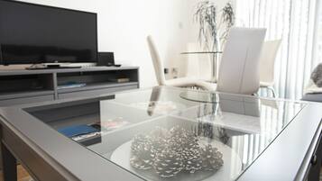 A modern living room with a glass coffee table, a flat-screen TV, a white chair, and a plant.