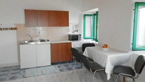 A compact kitchen with white cabinets, a microwave, and a sink. There is a dining area with a table and chairs, and a window with a green frame. - Villa Soti - Two-Bedroom Apartment with Sea View (Mlini)