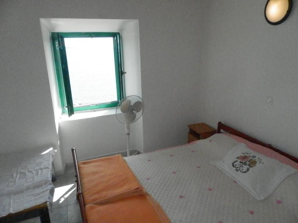 Una camera da letto con un letto, un comodino, una finestra con cornice verde e una ventola a parete.