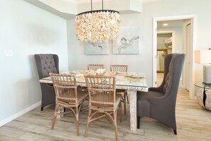 Un comedor con una mesa dispuesta para cuatro, rodeada de sillas de mimbre y un sillón oscuro. Un candelabro cuelga encima, y se exhiben obras de arte en la pared.